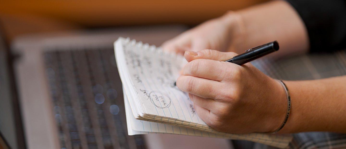 A person's hands writing on a notepad, held over an open laptop.